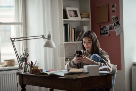 Téléchargez les photos : Jeune adolescente faisant ses devoirs et défilant smartphone dans sa chambre. - en image libre de droit