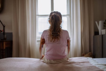 Photo for Rear view of little unhappy girl with broken leg sitting on a bed in her room. - Royalty Free Image