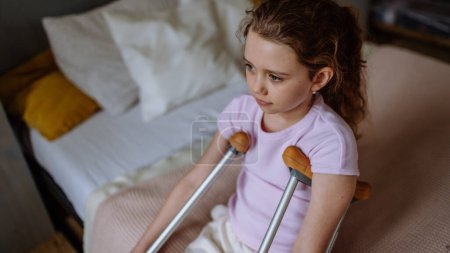 Téléchargez les photos : Petite fille malheureuse avec jambe cassée assise sur un lit dans sa chambre. - en image libre de droit