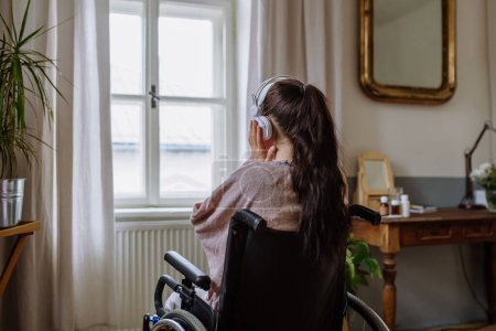Foto de Adolescente sentada en silla de ruedas, escuchando música y mirando por la ventana. - Imagen libre de derechos