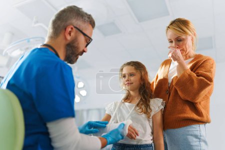 Téléchargez les photos : Mature médecin donnant gypse à la petite fille sur son bras cassé. - en image libre de droit