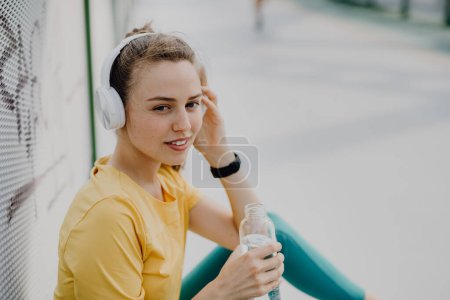 Foto de Mujer joven bebiendo agua y escuchando la música a través de auriculares, durante el trote en la ciudad, estilo de vida saludable y el concepto de deporte. - Imagen libre de derechos