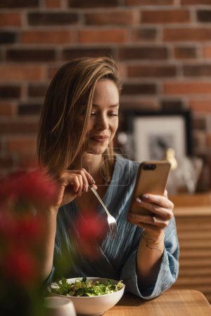 Photo for Young woman eating vegetable salad and scrolling her phone. - Royalty Free Image