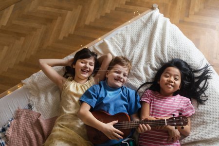 Foto de Vista superior de tres amigos felices acostados en la cama con ukelele. - Imagen libre de derechos
