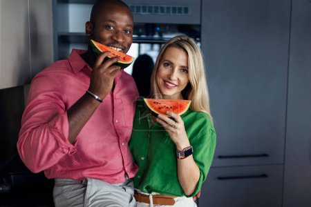 Téléchargez les photos : Couple multiracial mangeant un melon dans la cuisine pendant les chaudes journées ensoleillées. - en image libre de droit