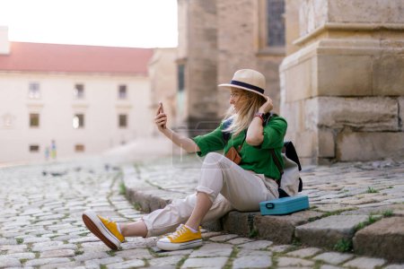 Téléchargez les photos : Jeune femme blonde voyage seule dans le vieux centre-ville, assise et utilisant un smartphone, prenant selfie. - en image libre de droit