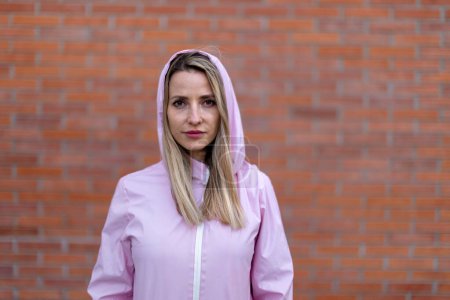Téléchargez les photos : Young blond woman with hood on head, standing in front of a brick wall. - en image libre de droit