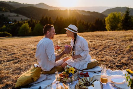 Foto de An attractive couple in love enjoying picnic and drinking wine on the hill at sunset. - Imagen libre de derechos
