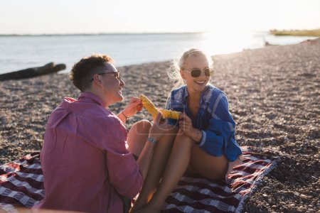 Foto de A happy young couple dating together in beach, sitting on a blanket and eating corn. Enjoying holiday time together. - Imagen libre de derechos