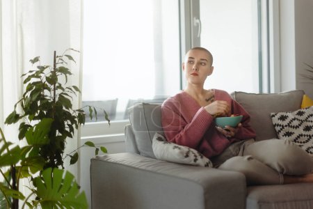 Photo for Young bald woman sitting on sofa and having a snack. - Royalty Free Image