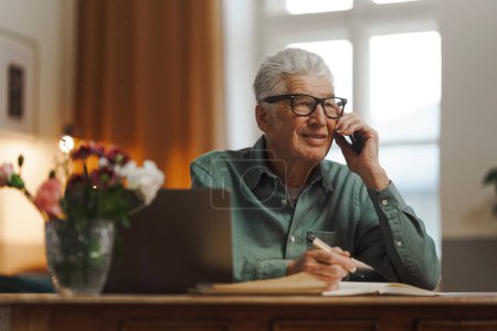 Téléchargez les photos : Homme âgé appelant et écrivant quelques notes dans son appartement. - en image libre de droit