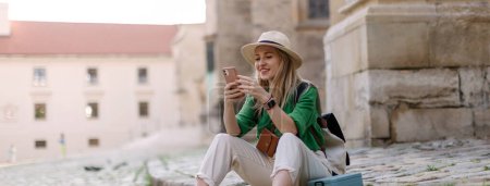 Foto de Young blond woman travel alone in old city centre, sitting and using smartphone. - Imagen libre de derechos