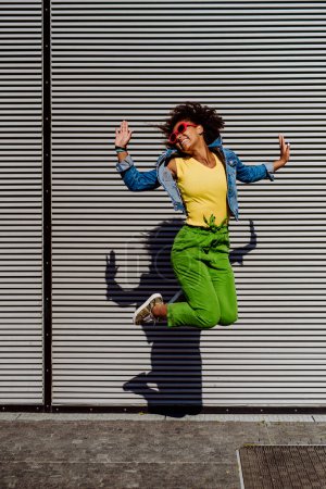 Foto de Young happy multiracial teenage girl with red sunglasses and afro hairstyle, jumping and posing outdoor. - Imagen libre de derechos