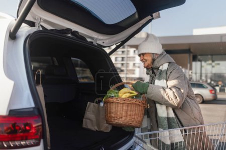 Foto de Mujer mayor dando su compra en el maletero del coche eléctrico. - Imagen libre de derechos