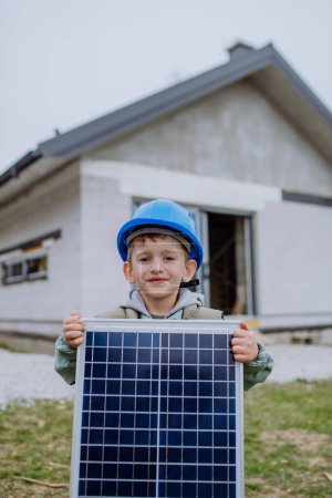 Téléchargez les photos : Portrait d'un petit garçon tenant un panneau solaire, devant leur nouvelle maison inachevée. - en image libre de droit