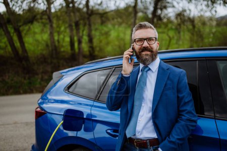 Téléchargez les photos : Homme d'affaires mature attendant de charger sa nouvelle voiture électrique. - en image libre de droit