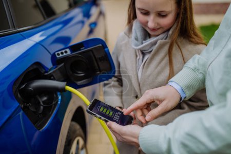 Photo for Close up of man and his daughter holding phone, checking charging of his electric car. - Royalty Free Image
