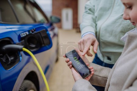 Photo for Close up of man and his daughter holding phone, checking charging of his electric car. - Royalty Free Image