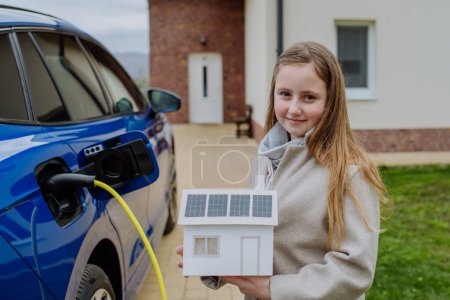 Foto de Niña sosteniendo el modelo de una casa de papel con paneles solares, esperando para cargar su coche eléctrico. - Imagen libre de derechos