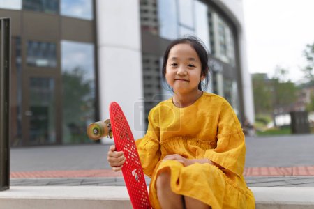 Foto de Feliz chica asiática con monopatín sentado al aire libre en la ciudad. - Imagen libre de derechos