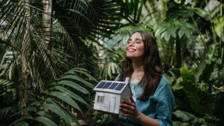 Foto de Mujer joven en selva sosteniendo modelo de casa con paneles solares, concepto de energía renovable y protección de la naturaleza. - Imagen libre de derechos