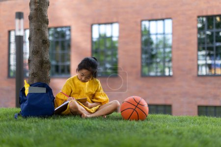Kleines asiatisches Mädchen sitzt im öffentlichen Park und schreibt ein paar Notizen. Sommerzeit.