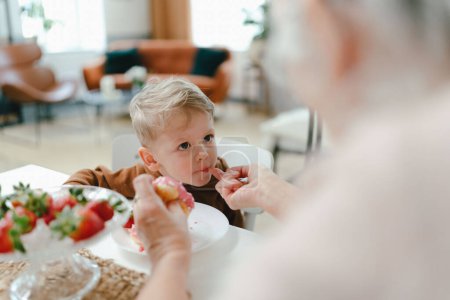 Foto de Un niño comiendo un bocadillo dulce con su abuela. Abuela alimentación gradson con donut. - Imagen libre de derechos