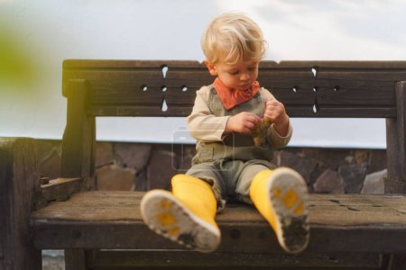 Foto de Niño en botas de goma amarillas sentado en un banco de madera y comiendo uvas de cosecha propia. - Imagen libre de derechos