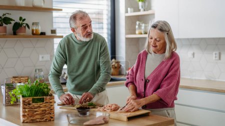Photo for Senior couple cooking together in the kitchen, preparing chicken for dinner. Spending quality time together. - Royalty Free Image