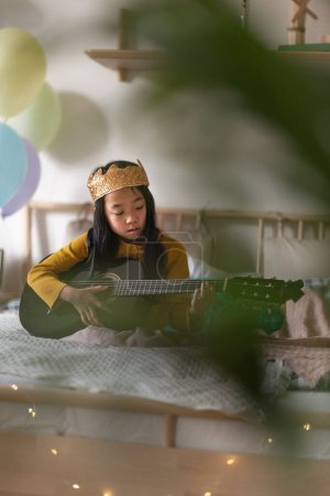 Téléchargez les photos : Petite princesse jouant de la guitare dans la pièce. Jeune fille indépendante apprenant à jouer de la guitare toute seule. - en image libre de droit