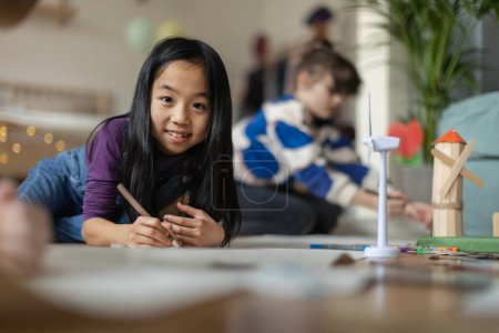 Photo for Portrait of a cute Asian girl working on her school project. Young girl lying on a floor and drawing. - Royalty Free Image