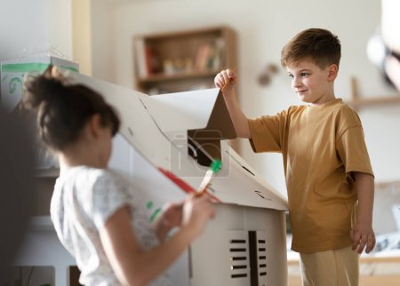 Téléchargez les photos : Heureux enfants peignant maison en carton fait à la main ensemble, travaillant sur le projet scolaire. Activités créatives d'intérieur pour enfants. - en image libre de droit