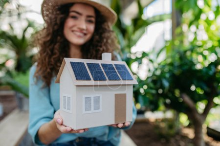 Foto de Mujer joven en un jardín botánico sosteniendo modelo de casa con paneles solares, concepto de energía verde. - Imagen libre de derechos