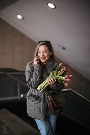 Photo for Young woman calling in a city with bouquet of tulips. - Royalty Free Image