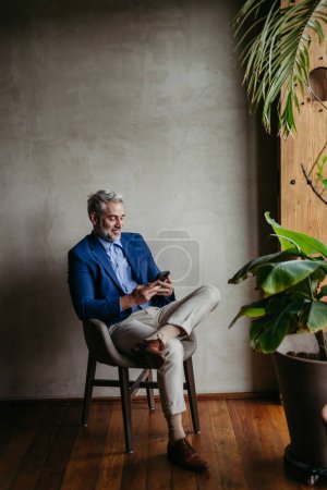 Téléchargez les photos : Portrait d'un homme d'affaires assis près de la fenêtre dans un bureau moderne plein de plantes. Homme dirigeant d'entreprise, exécutif, gestionnaire utilisant un téléphone intelligent.. - en image libre de droit