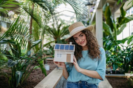 Photo for Young woman in a botanical garden holding model of house with solar panels, concept of green energy. - Royalty Free Image