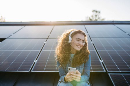 Photo for Portrait of young excited woman, owner on roof with solar panels, listening music. - Royalty Free Image