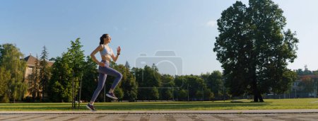 Foto de Vista lateral de la joven atlética en ropa deportiva corriendo en la ciudad por la mañana. Concepto de entrenamiento al aire libre. - Imagen libre de derechos