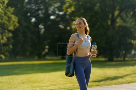 Téléchargez les photos : Plan moyen de gir athlétique en vêtements de sport avec tapis de yoga sur l'épaule et bouteille d'eau se dirigeant vers une séance d'entraînement pilates. - en image libre de droit