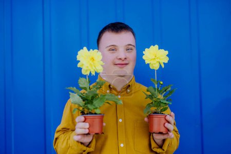 Photo for A happy young man with Down syndrome looking at camera and holding pot flowers against blue background. - Royalty Free Image