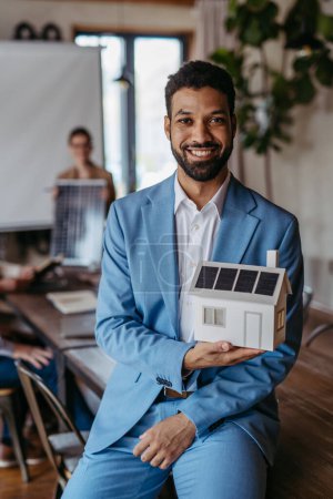 Téléchargez les photos : Portrait de jeune homme d'affaires confiant tenant modèle de maison avec panneaux solaires dans la salle de réunion. De jeunes collègues discutent de panneaux photovoltaïques, propres, verts, énergétiques. Entreprises utilisant des énergies renouvelables - en image libre de droit