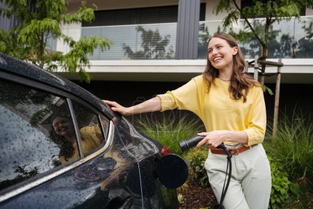 Foto de Mujer joven sosteniendo cable de alimentación de su coche, preparada para cargarlo en el hogar, concepto de transporte sostenible y económico. - Imagen libre de derechos