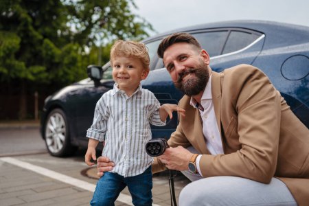 Téléchargez les photos : Gros plan de l'homme et de son petit fils tenant un câble d'alimentation de leur voiture électrique. - en image libre de droit
