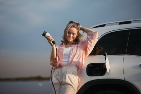 Foto de Mujer joven sosteniendo cable de alimentación de su coche, preparado para cargarlo, concepto de transporte sostenible y económico. - Imagen libre de derechos