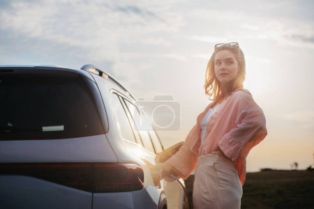 Photo for Young woman holding power supply cable from her car, prepared for charging it, sustainable and economic transportation concept. - Royalty Free Image