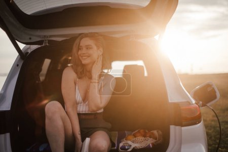 Téléchargez les photos : Jeune femme assise dans un coffre de voiture, attendant de le charger. - en image libre de droit