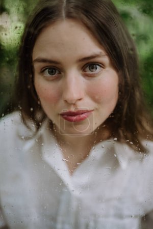 Photo for Portrait of a young woman in misty greenhouse through foggy glass with water droplets. - Royalty Free Image