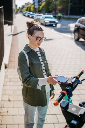 Téléchargez les photos : Jeune femme en ville prêt scooter avec une application. E-scooter système de partage. - en image libre de droit