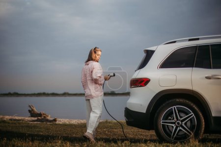 Téléchargez les photos : Jeune femme tenant le câble d'alimentation de sa voiture, prêt à le charger, concept de transport durable et économique. - en image libre de droit