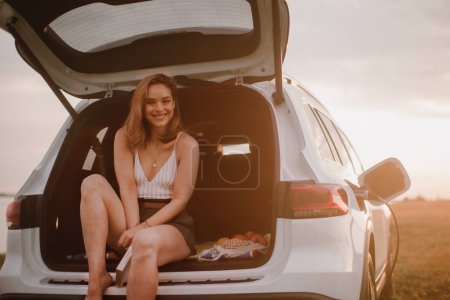 Téléchargez les photos : Jeune femme assise dans un coffre de voiture, attendant de le charger. - en image libre de droit